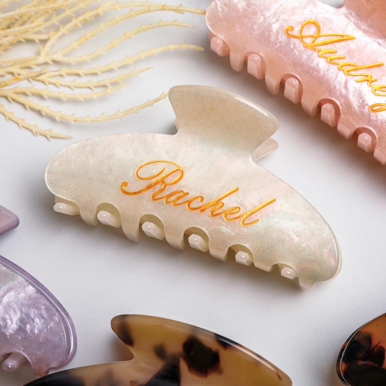 May include: Several personalised hair claw clips in different colours. The main clip is off-white with the name "Rachel" in gold script. Other clips include pink and tortoiseshell patterns, each with a name.