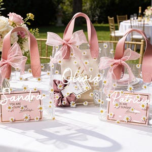 May include: Clear tote bags decorated with white daisies and pink accents. Each bag has a pink handle and a matching bow. The bags have personalized name cards that read "Sandra", "Olivia", and "Patricia".