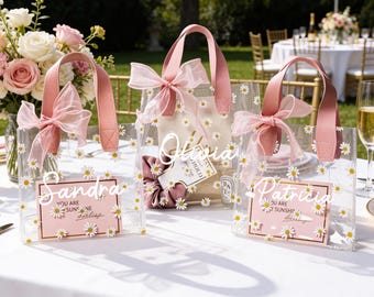 May include: Clear tote bags decorated with white daisies and pink accents. Each bag has a pink handle and a matching bow. The bags have personalized name cards that read "Sandra", "Olivia", and "Patricia".