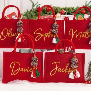 May include: Five red tote bags with gold script names: Olivia, Sophie, Matte, Daniel, and Jackson. Each bag has a matching handle and a decorative tassel with beads and tassels. Christmas-themed gifts are visible inside the bags.