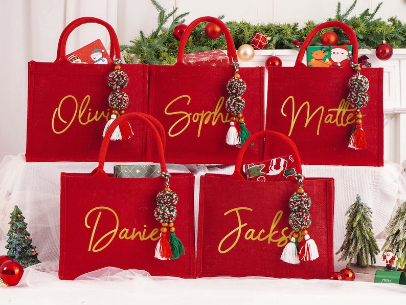 May include: Five red tote bags with gold script names: Olivia, Sophie, Matte, Daniel, and Jackson. Each bag has a matching handle and a decorative tassel with beads and tassels. Christmas-themed gifts are visible inside the bags.
