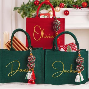 May include: Three Christmas-themed tote bags. A red bag with "Olivia" in gold script, and two green bags with "Daniel" and "Jackson" in gold script. Each bag has a decorative tassel. The bags are filled with gifts.