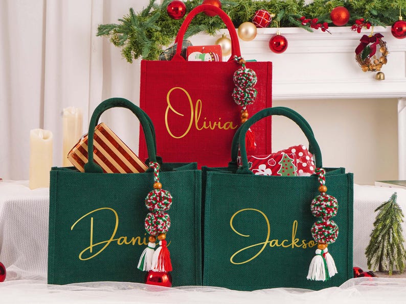 May include: Three Christmas-themed tote bags. A red bag with "Olivia" in gold script, and two green bags with "Daniel" and "Jackson" in gold script. Each bag has a decorative tassel. The bags are filled with gifts.