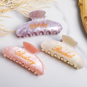 May include: Three pearlescent hair claw clips in pink, lavender, and cream. Each clip has a name in gold script: "Audrey", "Melody", and "Rachel". The clips are on a white marble surface with a sprig of dried flowers.