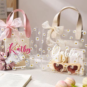 May include: Two clear tote bags decorated with white daisies. One bag has a pink handle and bow, the other has a cream handle and bow. Each bag has a name written on it, and contains items like sunglasses and scrunchies.