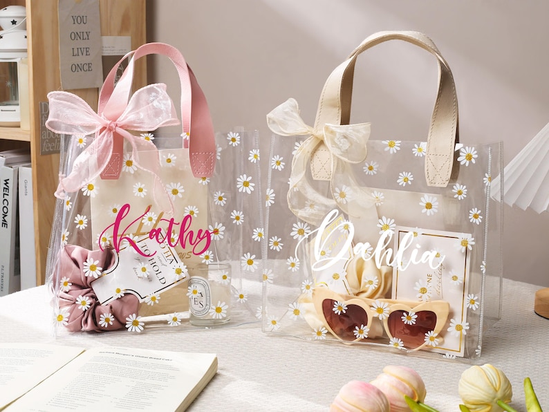 May include: Two clear tote bags decorated with white daisies. One bag has a pink handle and bow, the other has a cream handle and bow. Each bag has a name written on it, and contains items like sunglasses and scrunchies.
