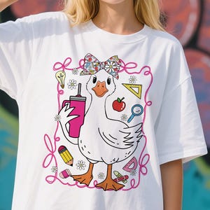 May include: White t-shirt featuring a cartoon goose wearing a bow and holding a pink tumbler. The goose is surrounded by school-themed illustrations, including a pencil, apple, and ruler. The design is outlined in pink.