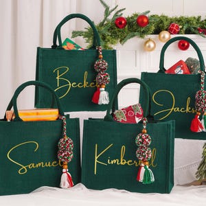 May include: Four green tote bags with gold script names: Beckett, Samuel, Jacks, and Kimberly. Each bag has a matching handle and a decorative tassel with red, white, and green accents. The bags are filled with gifts and are set against a festive holiday backdrop.