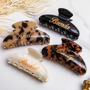 May include: Four personalised hair claw clips in different colours and patterns. One is black with "Brenda" in gold script, another is tortoiseshell with "Emily", a third is cream with "Rachel", and the last is tortoiseshell with "Amelia".