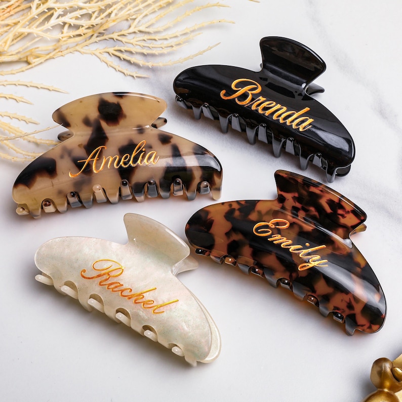 May include: Four personalised hair claw clips in different colours and patterns. One is black with "Brenda" in gold script, another is tortoiseshell with "Emily", a third is cream with "Rachel", and the last is tortoiseshell with "Amelia".