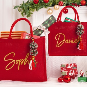 May include: Two red tote bags with gold lettering, one reading "Sophie" and the other "Daniel." Each bag has a matching handle and a decorative tassel. The bags are filled with gifts and are decorated with Christmas ornaments.