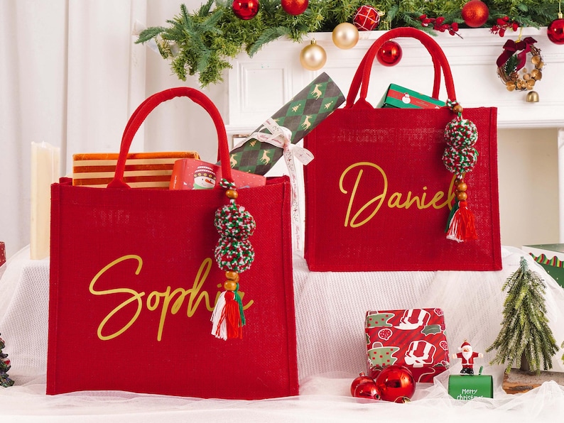 May include: Two red tote bags with gold lettering, one reading "Sophie" and the other "Daniel." Each bag has a matching handle and a decorative tassel. The bags are filled with gifts and are decorated with Christmas ornaments.