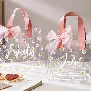 May include: Two clear tote bags with pink handles and bows, decorated with white daisies. One bag says "Emily" and the other says "Julia" in white script. Heart-shaped sunglasses and a book are in the foreground.