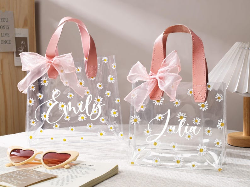 May include: Two clear tote bags with pink handles and bows, decorated with white daisies. One bag says "Emily" and the other says "Julia" in white script. Heart-shaped sunglasses and a book are in the foreground.