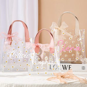 May include: Three clear tote bags with daisy patterns and personalized names in cursive. Each bag has a pink handle and a matching bow. The bags are on a white surface with a vase of flowers in the background.