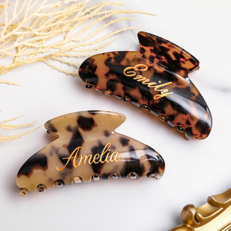 May include: Two tortoiseshell hair claw clips with gold lettering. One clip says "Emily" and the other says "Amelia". The clips are brown and tan with a glossy finish. The hair accessories are on a white surface.