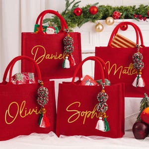 May include: Four red tote bags with gold script names: Daniel, Olivia, Sophie, and Matteo. Each bag has a matching red handle and a decorative tassel. The bags are filled with gifts, and are decorated for Christmas.