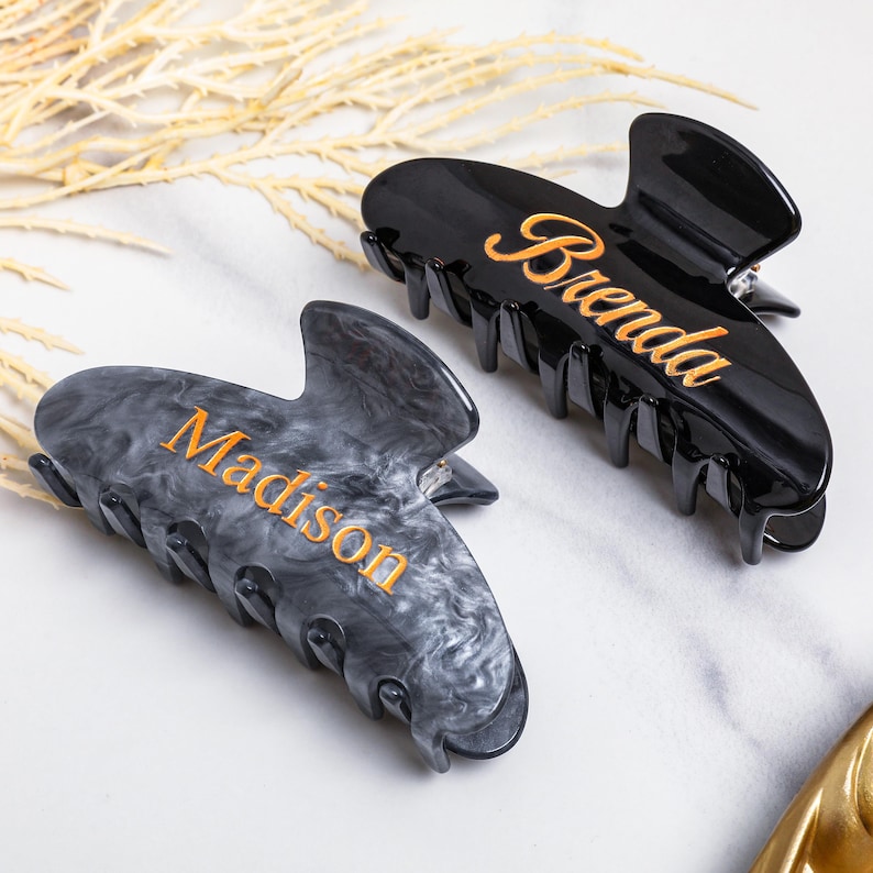 May include: Two personalised hair claw clips. One is black with "Brenda" in gold script, the other is grey marble with "Madison" in gold script. The clips are on a white surface with dried decorative plants.