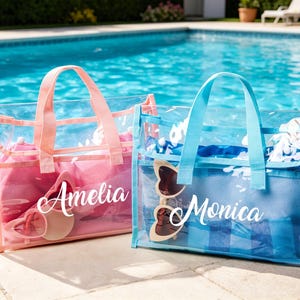 May include: Two clear tote bags with mesh pockets and matching handles sit poolside. One is pink and the other is blue. The pink bag has the name "Amelia" in white script, and the blue bag has the name "Monica". Both bags contain items like sunglasses and towels.