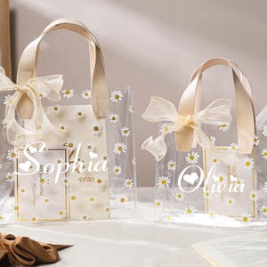 May include: Two clear tote bags with daisy patterns and beige handles. One bag says "Sophia" and the other says "Olivia" in white script. Both bags have a sheer bow at the top. The bags are on a white surface.