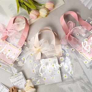 May include: Clear tote bags with daisy patterns, pink handles, and bows. Each bag has a pink tag with a name written in cursive. The bags are decorated with small white daisies.