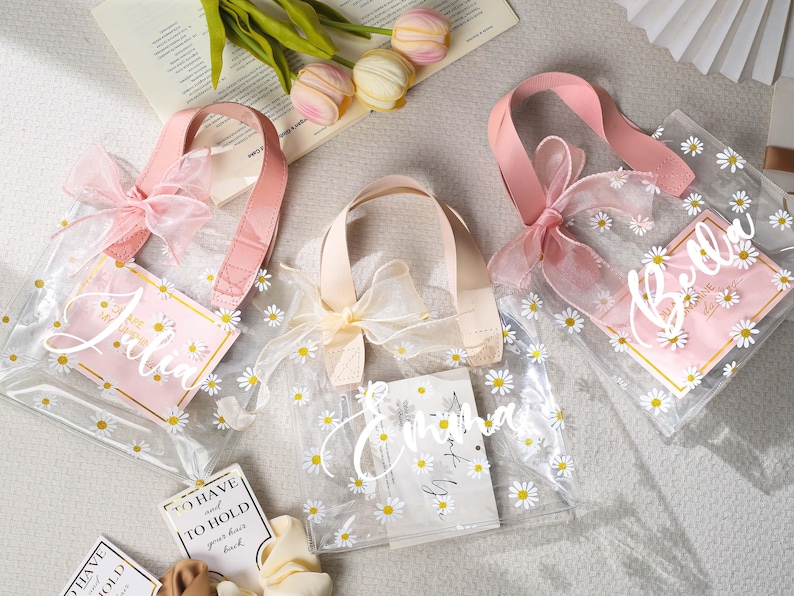 May include: Clear tote bags with daisy patterns, pink handles, and bows. Each bag has a pink tag with a name written in cursive. The bags are decorated with small white daisies.