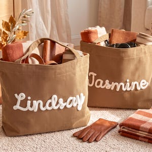 Könnte beinhalten: Zwei beigefarbene Cord-Einkaufstaschen mit weiß gestickten Namen. Eine Tasche trägt den Namen Lindsay und die andere den Namen Jasmine. Beide Taschen sind mit verschiedenen Gegenständen gefüllt, darunter gefaltete Textilien und kleine Accessoires.