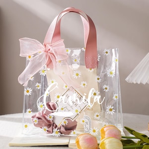May include: Clear tote bag with daisy print and pink leather handles. The bag is decorated with a pink bow and contains a scrunchie, a small candle, and a card. The text "Camila" is written on the front of the bag.
