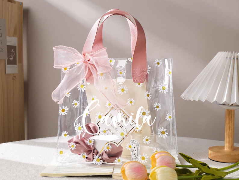May include: Clear tote bag with daisy print and pink leather handles. The bag is decorated with a pink bow and contains a scrunchie, a small candle, and a card. The text "Camila" is written on the front of the bag.