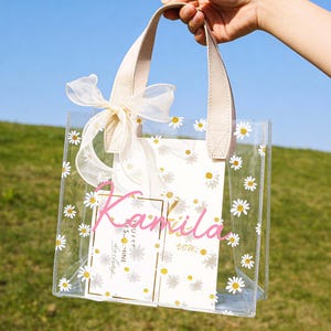 May include: Clear tote bag decorated with white daisies and a cream-colored bow. The bag has cream-colored handles and contains a card with the name "Kamila" in pink script. The background is a grassy hill and blue sky.