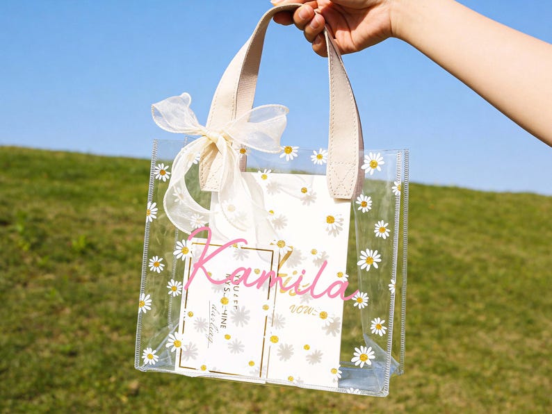 May include: Clear tote bag decorated with white daisies and a cream-colored bow. The bag has cream-colored handles and contains a card with the name "Kamila" in pink script. The background is a grassy hill and blue sky.