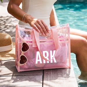May include: A rectangular clear plastic tote bag with light pink trim and handles. The front features the white text ARK printed in a bold sans-serif font. A pair of light pink heart-shaped sunglasses is attached to the side of the bag.