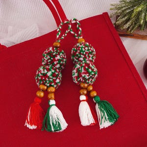 May include: Two decorative Christmas ornaments with red, white, and green yarn pom-poms, wooden beads, and tassels. The ornaments are hanging over a red tote bag. A red apple is in the background.