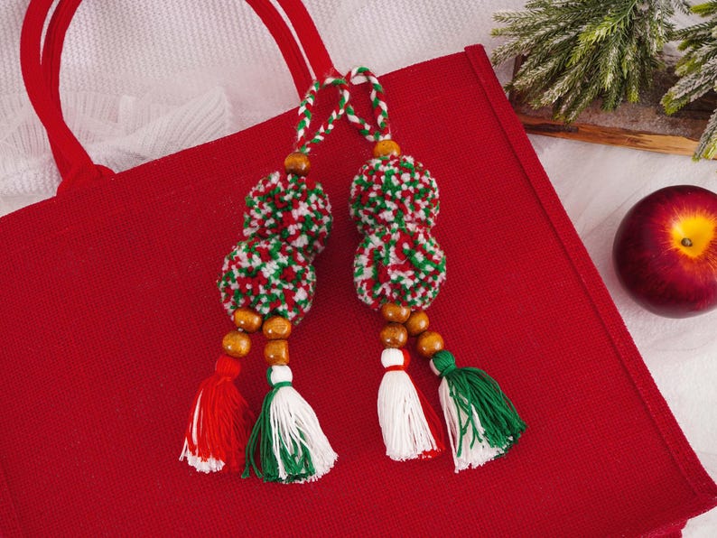 May include: Two decorative Christmas ornaments with red, white, and green yarn pom-poms, wooden beads, and tassels. The ornaments are hanging over a red tote bag. A red apple is in the background.