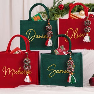 May include: Four Christmas-themed tote bags in red and green, each with a name in gold script. The bags have matching handles and decorative tassels. Some bags contain small gift boxes. Festive holiday decor is in the background.
