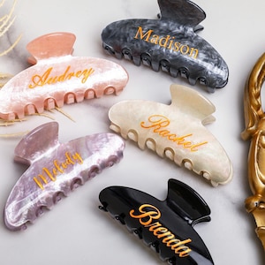 May include: Several personalised hair claw clips in shades of pink, grey, black, and white. Each clip has a name in gold script: Audrey, Madison, Rachel, Melody, and Brenda. The clips are made of a marbled material.