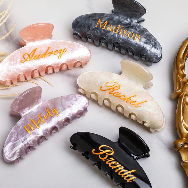 May include: Several personalised hair claw clips in shades of pink, grey, black, and white. Each clip has a name in gold script: Audrey, Madison, Rachel, Melody, and Brenda. The clips are made of a marbled material.