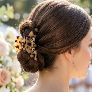 May include: A tortoiseshell hair claw clip, inscribed with the name "Amelia" in gold lettering, securing a woman's brown hair in a bun. The background is blurred, with flowers and greenery visible.