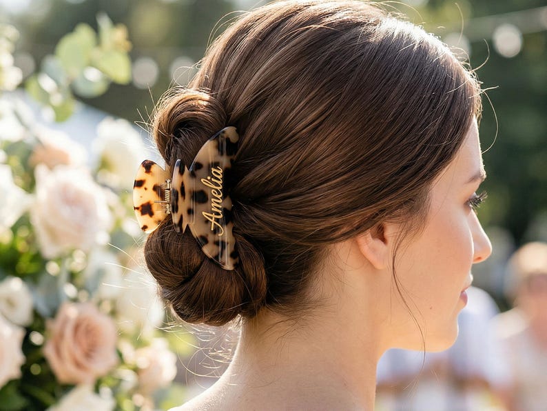 May include: A tortoiseshell hair claw clip, inscribed with the name "Amelia" in gold lettering, securing a woman's brown hair in a bun. The background is blurred, with flowers and greenery visible.