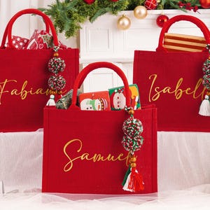 May include: Three red tote bags with gold lettering, each bearing a name: Fabian, Samuel, and Isabel. The bags have matching handles and decorative tassels. The bags are filled with gifts, and are decorated for Christmas.