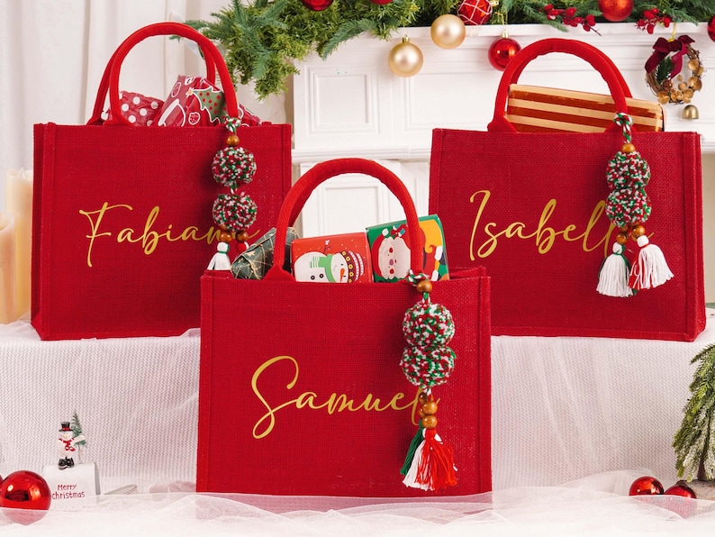 May include: Three red tote bags with gold lettering, each bearing a name: Fabian, Samuel, and Isabel. The bags have matching handles and decorative tassels. The bags are filled with gifts, and are decorated for Christmas.
