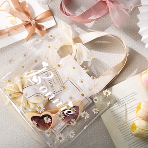 May include: A clear tote bag with daisy accents, filled with gifts, including heart-shaped sunglasses and a bow. The bag has a cream-colored handle and is decorated with small white daisies. A gift card with a ribbon and a book with tulips are also visible.