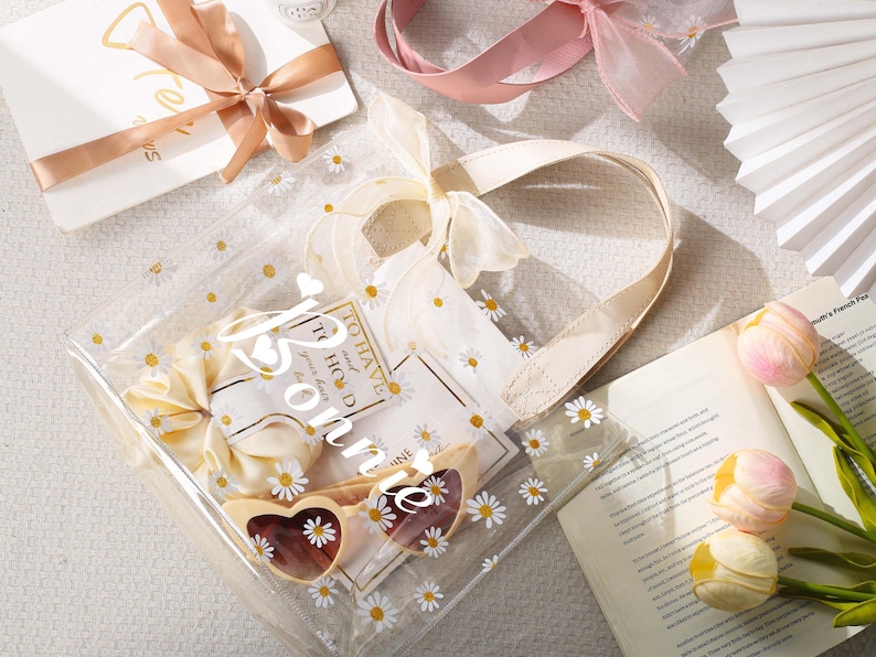 May include: A clear tote bag with daisy accents, filled with gifts, including heart-shaped sunglasses and a bow. The bag has a cream-colored handle and is decorated with small white daisies. A gift card with a ribbon and a book with tulips are also visible.