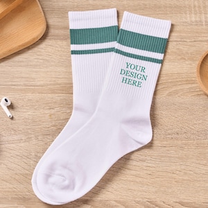 Può includere: Un paio di calzini bianchi con due strisce verdi nella parte superiore. Il testo "YOUR DESIGN HERE" è stampato in verde su un calzino. I calzini sono esposti su una superficie di legno.