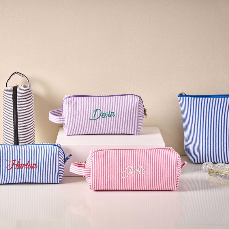 Pode incluir: Uma cole&ccedil;&atilde;o de bolsas de cosm&eacute;ticos listradas em v&aacute;rias cores, incluindo rosa, roxo e azul. Cada bolsa apresenta um nome bordado na frente em uma cor contrastante. As bolsas t&ecirc;m fecho de z&iacute;per e uma pequena al&ccedil;a.