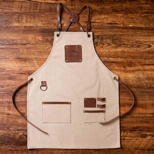Op de afbeelding: Een beige canvas schort met bruine leren banden, een bruine leren borstpatch met de naam Alison en een bergafbeelding, een metalen ring en meerdere bruine leren zakken en lussen voor opslag, vastgezet met koperkleurige metalen klinknagels.