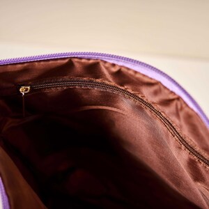 Pode incluir: Close-up de uma bolsa roxa com z&iacute;per e interior marrom. O z&iacute;per &eacute; marrom escuro com um puxador dourado. O interior da bolsa &eacute; de cor marrom s&oacute;lida.