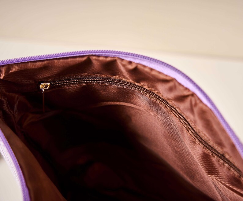 Pode incluir: Close-up de uma bolsa roxa com z&iacute;per e interior marrom. O z&iacute;per &eacute; marrom escuro com um puxador dourado. O interior da bolsa &eacute; de cor marrom s&oacute;lida.