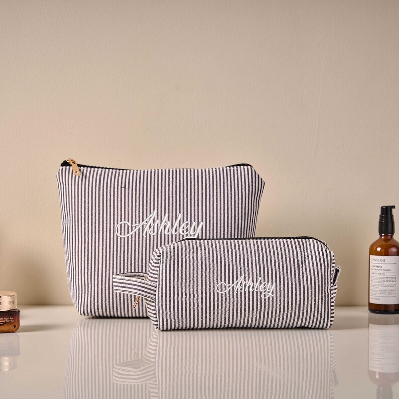 Pode incluir: Duas bolsas de cosm&eacute;ticos listradas em preto e branco, uma grande e outra pequena. O nome "Ashley" &eacute; bordado em branco em cada bolsa. As bolsas t&ecirc;m z&iacute;peres pretos e a menor tem uma pequena al&ccedil;a. O fundo &eacute; de cor neutra.