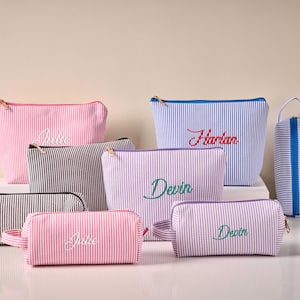 Pode incluir: Cole&ccedil;&atilde;o de n&eacute;cessaires personalizados em v&aacute;rios tamanhos e cores, incluindo rosa, azul, preto e lavanda. Cada bolsa apresenta um padr&atilde;o de listras seersucker e nomes bordados em escrita cursiva. Z&iacute;peres dourados e uma pequena al&ccedil;a vis&iacute;veis.
